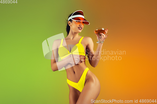 Image of Fashion portrait of young fit and sportive woman with cocktail in stylish yellow luxury swimwear on gradient background. Perfect body ready for summertime.