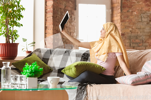 Image of A pretty young muslim woman at home during quarantine and self-insulation, using tablet for selfie or videocall, online lessons