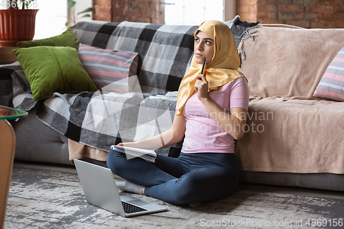 Image of A pretty young muslim woman at home during quarantine and self-insulation, using laptop, listen to music, watching cinema, serials, shopping, studying