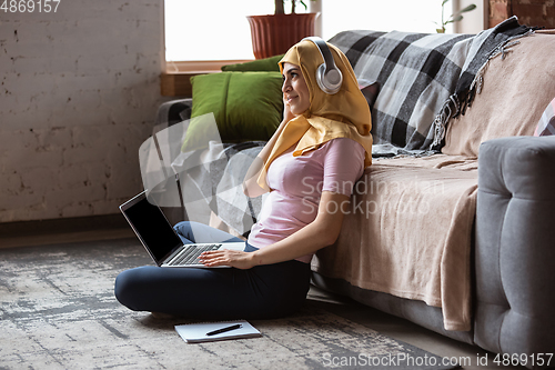 Image of A pretty young muslim woman at home during quarantine and self-insulation, using laptop, listen to music, watching cinema, serials, shopping, studying