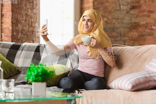 Image of A pretty young muslim woman at home during quarantine and self-insulation, using smartphone for selfie or videocall, online lessons, shopping