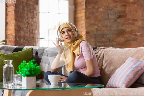 Image of A pretty young muslim woman at home during quarantine and self-insulation, using headphones, listen to music, enjoying