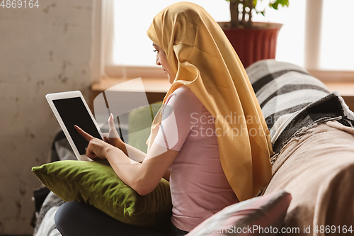 Image of A pretty young muslim woman at home during quarantine and self-insulation, using tablet for selfie or videocall, online lessons