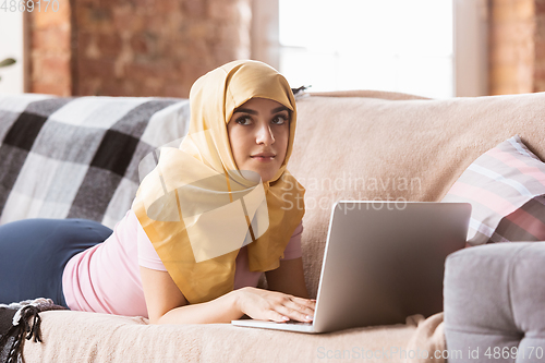 Image of A pretty young muslim woman at home during quarantine and self-insulation, using laptop, listen to music, watching cinema, serials, shopping, studying