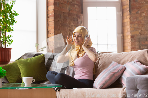 Image of A pretty young muslim woman at home during quarantine and self-insulation, using headphones, listen to music, enjoying