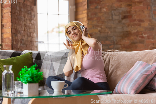 Image of A pretty young muslim woman at home during quarantine and self-insulation, using headphones, listen to music, enjoying