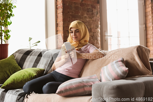 Image of A pretty young muslim woman at home during quarantine and self-insulation, using smartphone for selfie or videocall, online lessons, shopping