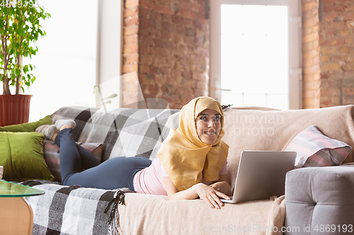 Image of A pretty young muslim woman at home during quarantine and self-insulation, using laptop, listen to music, watching cinema, serials, shopping, studying