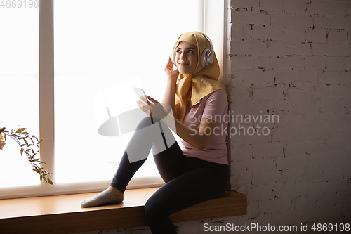 Image of A pretty young muslim woman at home during quarantine and self-insulation, using smartphone for selfie or videocall, online lessons, shopping