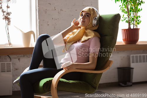 Image of A pretty young muslim woman at home during quarantine and self-insulation, using headphones, listen to music, enjoying