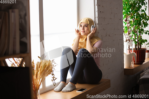 Image of A pretty young muslim woman at home during quarantine and self-insulation, using headphones, listen to music, enjoying