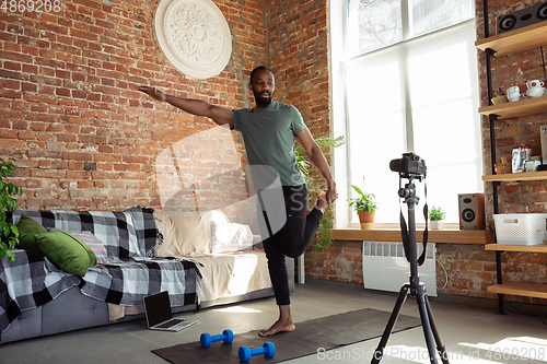 Image of Young african-american man teaching at home online courses of fitness, aerobic, sporty lifestyle during quarantine, reording on camera, streaming