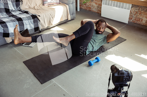 Image of Young african-american man teaching at home online courses of fitness, aerobic, sporty lifestyle during quarantine, reording on camera, streaming