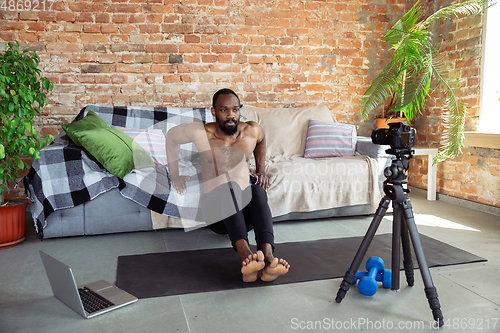 Image of Young african-american man teaching at home online courses of fitness, aerobic, sporty lifestyle during quarantine, reording on camera, streaming