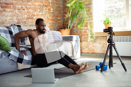 Image of Young african-american man teaching at home online courses of fitness, aerobic, sporty lifestyle during quarantine, reording on camera, streaming