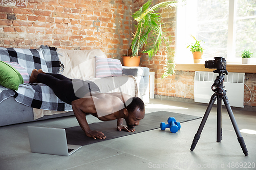 Image of Young african-american man teaching at home online courses of fitness, aerobic, sporty lifestyle during quarantine, reording on camera, streaming