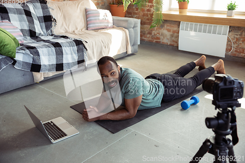 Image of Young african-american man teaching at home online courses of fitness, aerobic, sporty lifestyle during quarantine, reording on camera, streaming