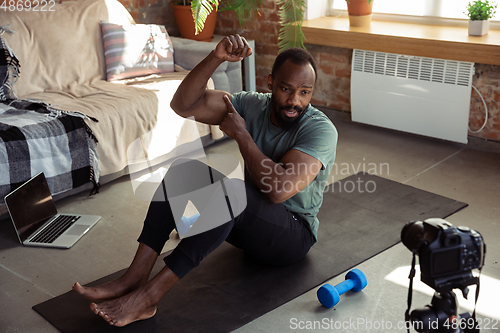 Image of Young african-american man teaching at home online courses of fitness, aerobic, sporty lifestyle during quarantine, reording on camera, streaming