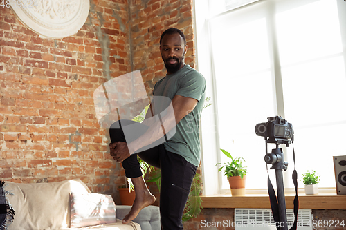 Image of Young african-american man teaching at home online courses of fitness, aerobic, sporty lifestyle during quarantine, reording on camera, streaming