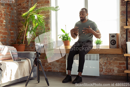 Image of Young african-american man teaching at home online courses of fitness, aerobic, sporty lifestyle during quarantine, reording on camera, streaming
