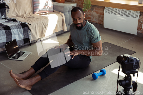 Image of Young african-american man teaching at home online courses of fitness, aerobic, sporty lifestyle during quarantine, reording on camera, streaming