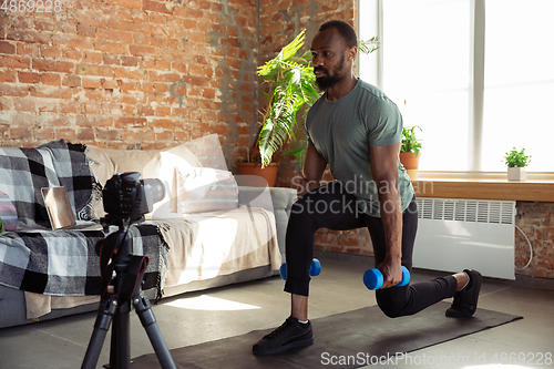 Image of Young african-american man teaching at home online courses of fitness, aerobic, sporty lifestyle during quarantine, reording on camera, streaming