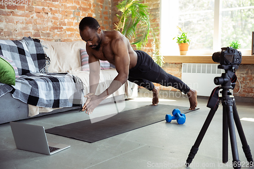 Image of Young african-american man teaching at home online courses of fitness, aerobic, sporty lifestyle during quarantine, reording on camera, streaming