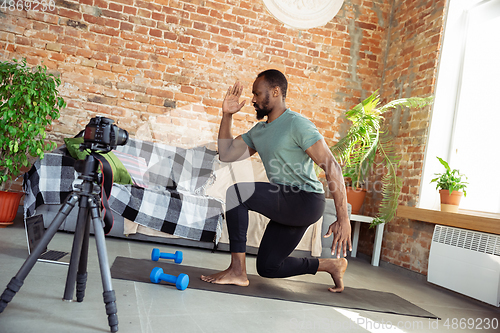 Image of Young african-american man teaching at home online courses of fitness, aerobic, sporty lifestyle during quarantine, reording on camera, streaming