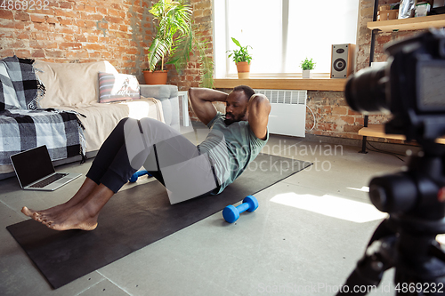 Image of Young african-american man teaching at home online courses of fitness, aerobic, sporty lifestyle during quarantine, reording on camera, streaming