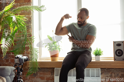 Image of Young african-american man teaching at home online courses of fitness, aerobic, sporty lifestyle during quarantine, reording on camera, streaming