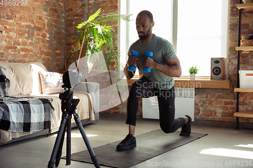 Image of Young african-american man teaching at home online courses of fitness, aerobic, sporty lifestyle during quarantine, reording on camera, streaming