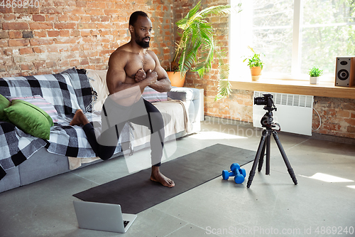 Image of Young african-american man teaching at home online courses of fitness, aerobic, sporty lifestyle during quarantine, reording on camera, streaming