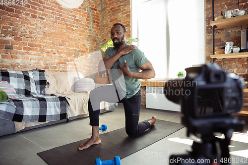 Image of Young african-american man teaching at home online courses of fitness, aerobic, sporty lifestyle during quarantine, reording on camera, streaming