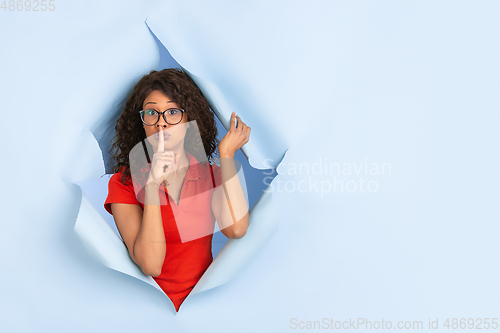 Image of Cheerful young woman poses in torn blue paper hole background, emotional and expressive