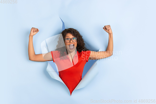 Image of Cheerful young woman poses in torn blue paper hole background, emotional and expressive