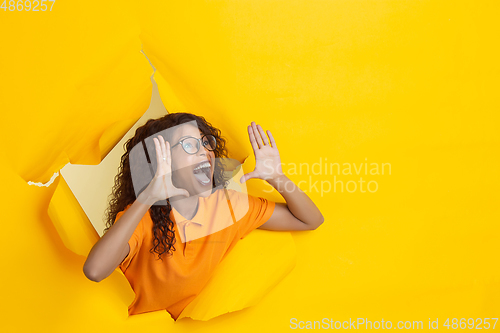 Image of Cheerful young woman poses in torn yellow paper hole background, emotional and expressive