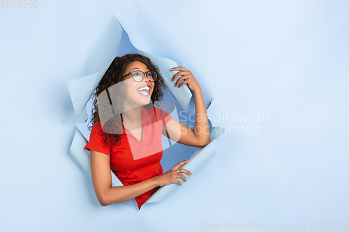 Image of Cheerful young woman poses in torn blue paper hole background, emotional and expressive