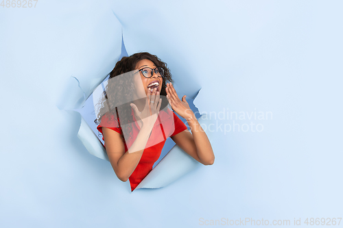 Image of Cheerful young woman poses in torn blue paper hole background, emotional and expressive