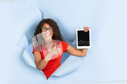 Image of Cheerful young woman poses in torn blue paper hole background, emotional and expressive