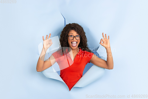 Image of Cheerful young woman poses in torn blue paper hole background, emotional and expressive