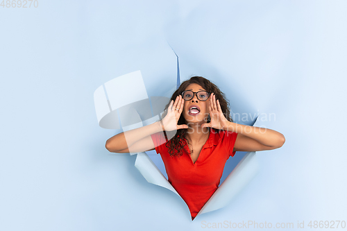 Image of Cheerful young woman poses in torn blue paper hole background, emotional and expressive