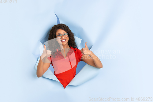 Image of Cheerful young woman poses in torn blue paper hole background, emotional and expressive