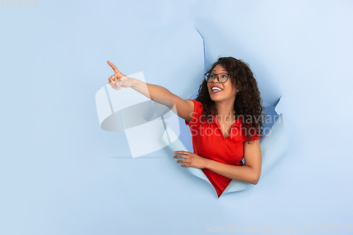 Image of Cheerful young woman poses in torn blue paper hole background, emotional and expressive