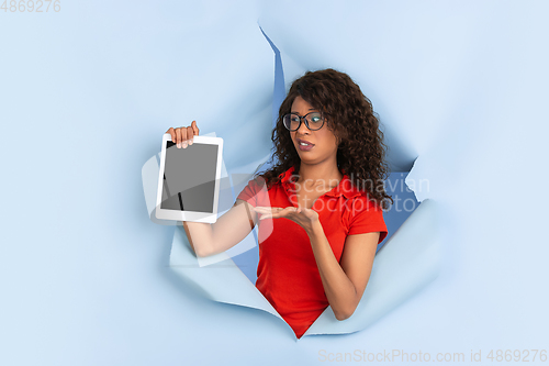 Image of Cheerful young woman poses in torn blue paper hole background, emotional and expressive