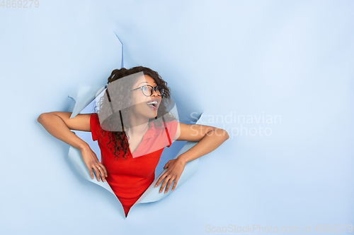 Image of Cheerful young woman poses in torn blue paper hole background, emotional and expressive