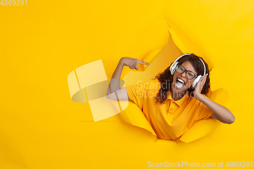 Image of Cheerful young woman poses in torn yellow paper hole background, emotional and expressive