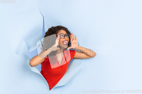 Image of Cheerful young woman poses in torn blue paper hole background, emotional and expressive