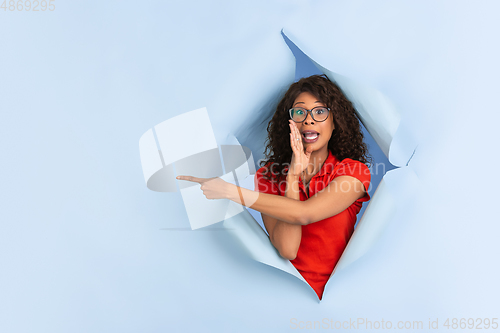 Image of Cheerful young woman poses in torn blue paper hole background, emotional and expressive