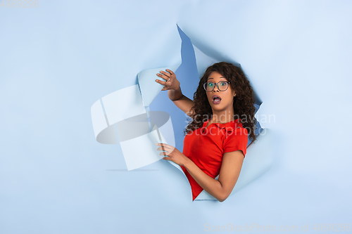 Image of Cheerful young woman poses in torn blue paper hole background, emotional and expressive