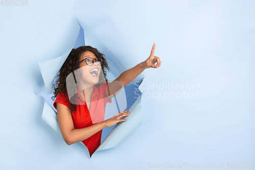 Image of Cheerful young woman poses in torn blue paper hole background, emotional and expressive